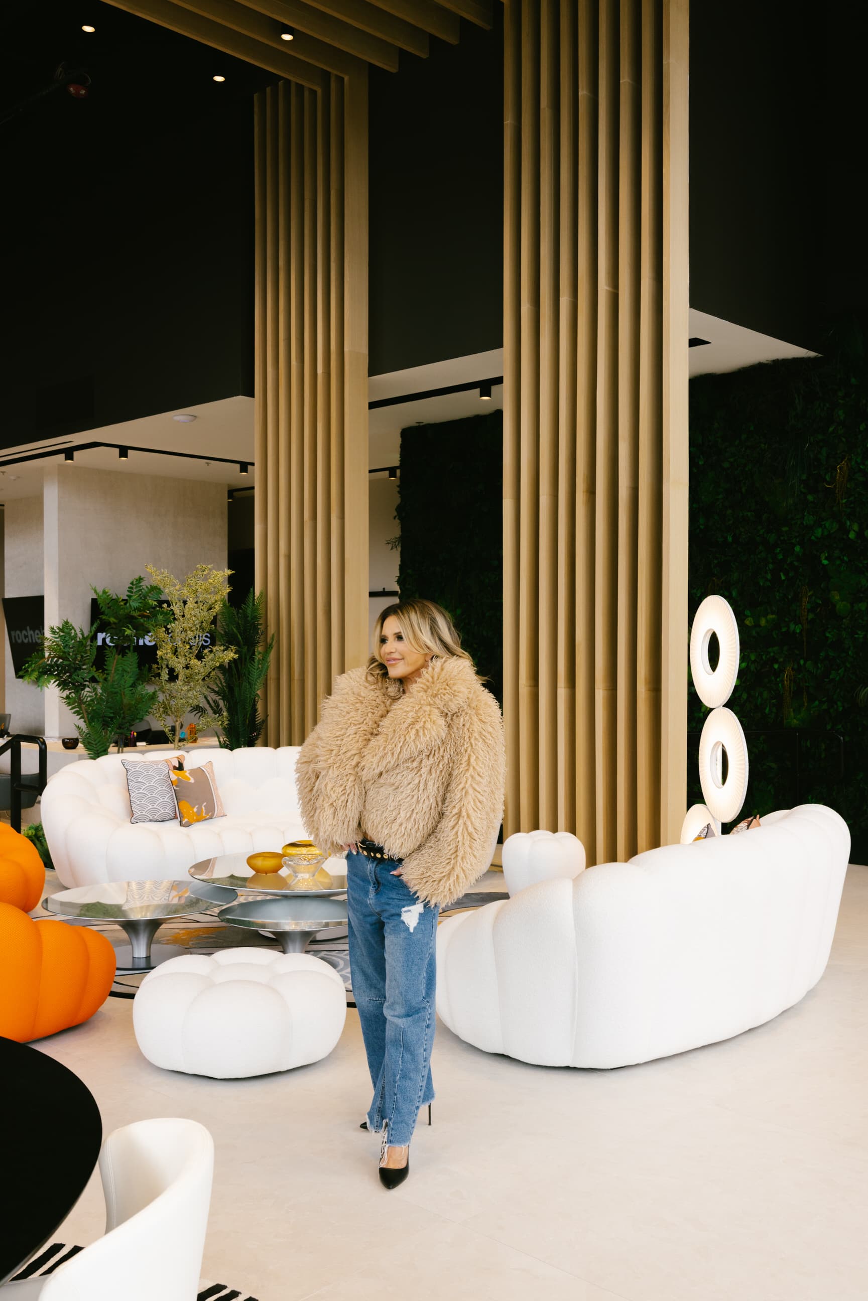 Giselle Koy in a tan shearling coat among ivory bubble sofas and bamboo columns in a designed showroom
