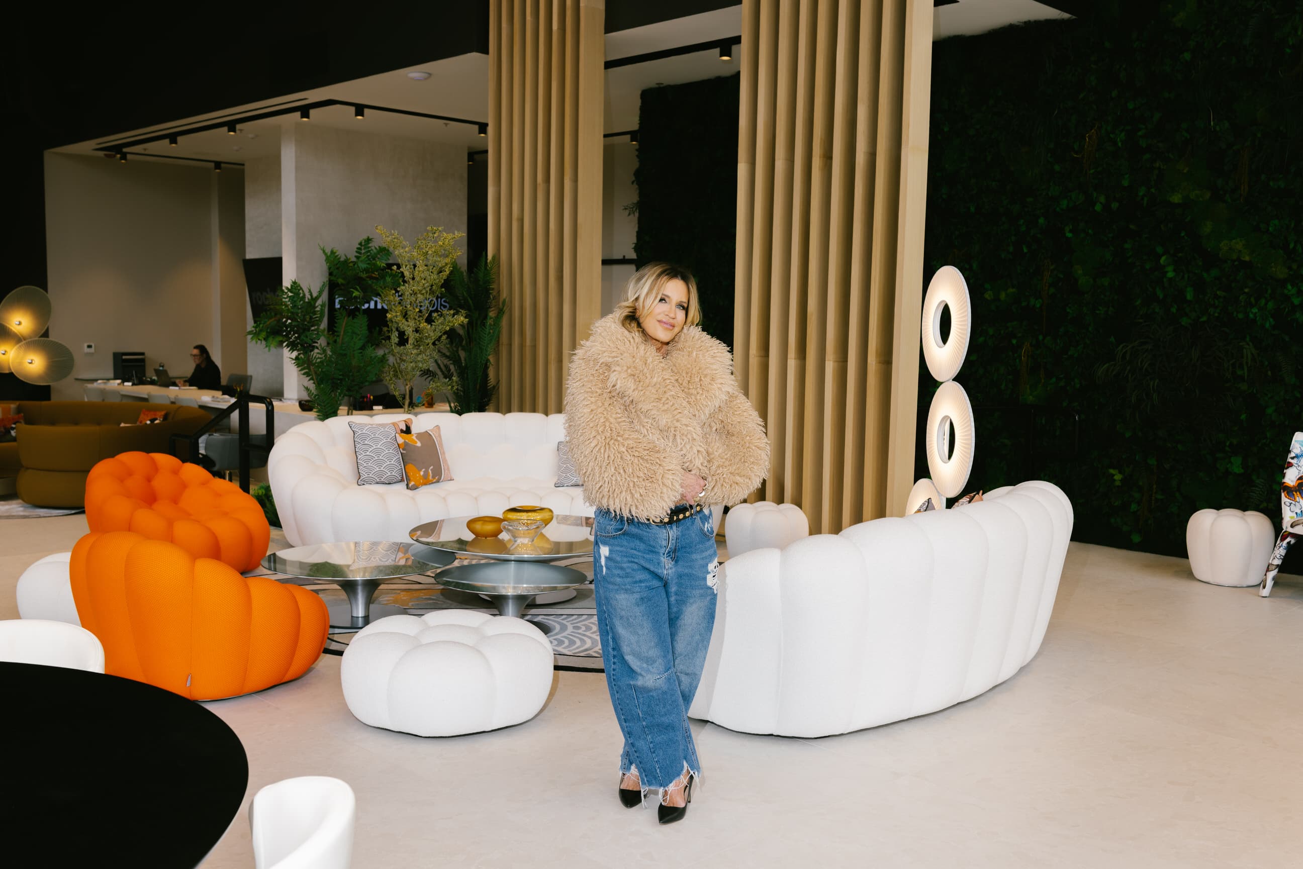 Giselle Koy in a tan shearling coat and ripped jeans, standing full-length among ivory bouclé bubble sofas, bamboo columns, and a living moss wall in a designed showroom