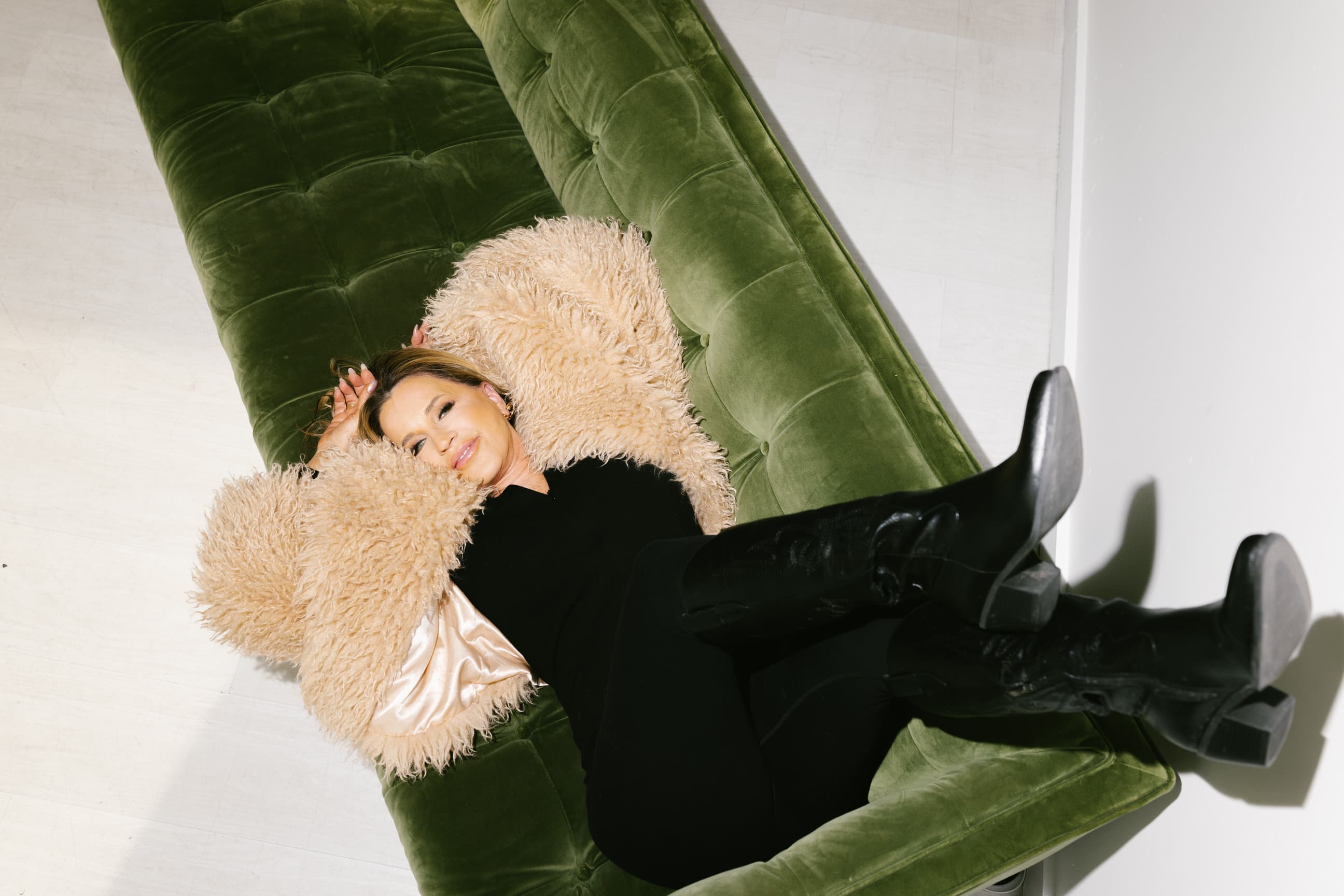 Giselle Koy seated on the floor in front of a green velvet chesterfield sofa, fur coat draped, looking calmly off-camera
