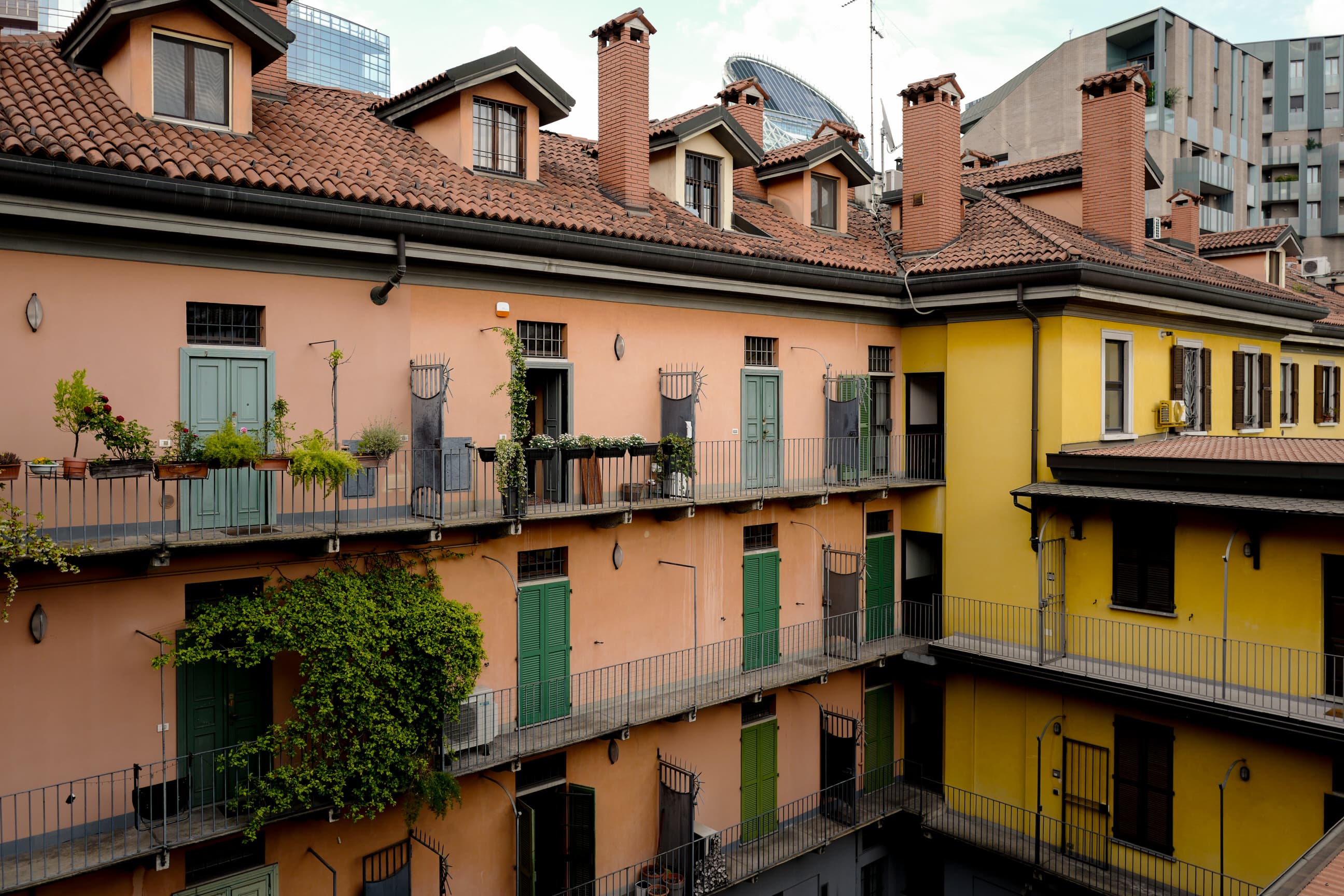 Milan courtyard — terracotta and yellow buildings with green shutters, balconies draped in vines, modern glass towers rising behind